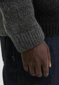 Dunkelhäutige Hand, die seitlich ruht, trägt einen dicken grauen Strickpullover und dunkellblaue Jeans.
