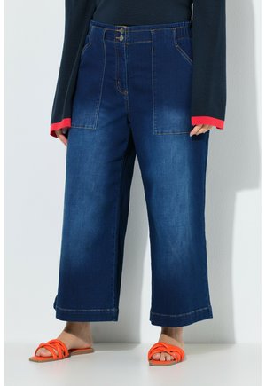 Luźne, szerokie spodnie z ciemnoniebieskiego denimu z przednimi naszywanymi kieszeniami, zestawione z jasnopomarańczowymi, płaskimi sandałami na paski oraz długim czarnym topem z czerwonymi mankietami.