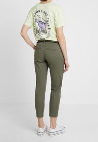 T-shirt vert clair avec un motif et du texte violets au dos, associé à un pantalon olive ajusté et des baskets blanches.