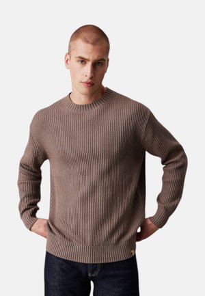 Giovane uomo con i capelli corti che indossa un maglione a coste marrone chiaro e jeans scuri, in piedi con le mani sui fianchi davanti a uno sfondo bianco.