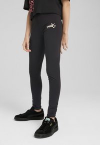 Černé legíny vyrobené z elastického materiálu, s zlatým logem Puma na stehně. Spárovány s černými semišovými teniskami.