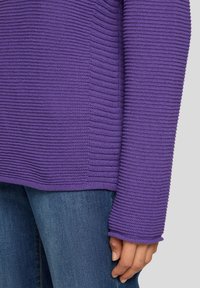 Lila gerippter Pullover aus strukturiertem Material mit langen Ärmeln und breiten Bündchen, getragen mit blauen Jeans. Nahaufnahme des Ärmel- und Saumdetails.