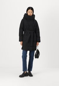 Abrigo de plumas negro con cuello alto y cinturón, con una superficie texturizada. Combinado con jeans azules y zapatos de plataforma de cuero negros.
