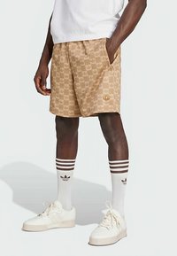 Calções castanhos claros com padrão xadrez texturizado e um discreto logótipo de coroa. Combinados com meias brancas com riscas castanhas e detalhes da marca, e ténis brancos.