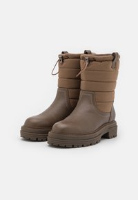 Bottes imperméables marron avec une partie inférieure en cuir et une partie supérieure matelassée. Dotées d'une semelle en caoutchouc texturé et d'une fermeture à cordon réglable.