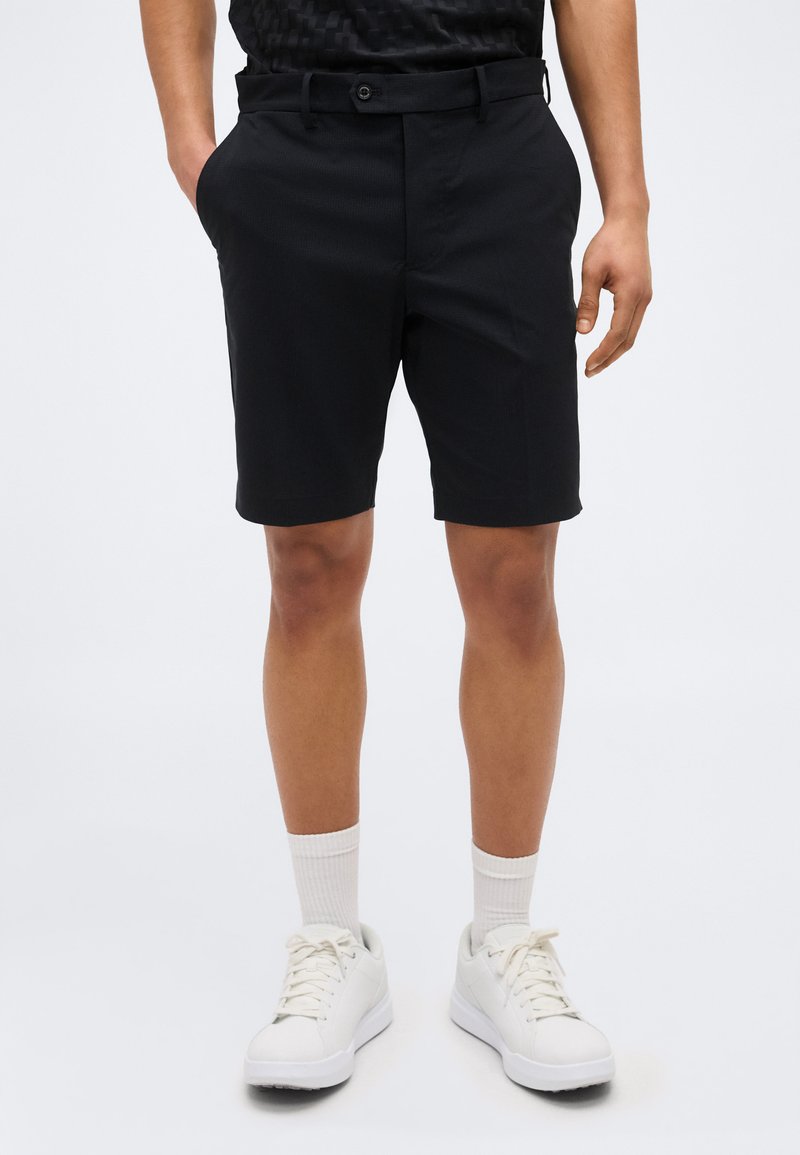 Άνδρας που φοράει μαύρα, κομμένα κομψά σορτς, λευκές κάλτσες μέχρι τον αστράγαλο και λευκά αθλητικά παπούτσια με κορδόνια, στέκεται με τα χέρια στις τσέπες.