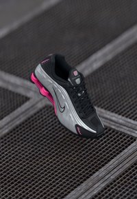 Czarno-srebrne buty do biegania Nike z różowymi akcentami zawieszone nad metalową siatką, podkreślające smukły design i amortyzację.