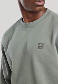 Felpa in cotone verde con scollatura tonda, caratterizzata da un piccolo logo quadrato applicato sul petto e polsini a coste. Texture morbida, vestibilità casual.