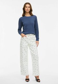 Le mannequin se tient avec une main dans sa poche, portant un pull bleu texturé et un pantalon large à fleurs blanches, ainsi que des talons hauts noirs.