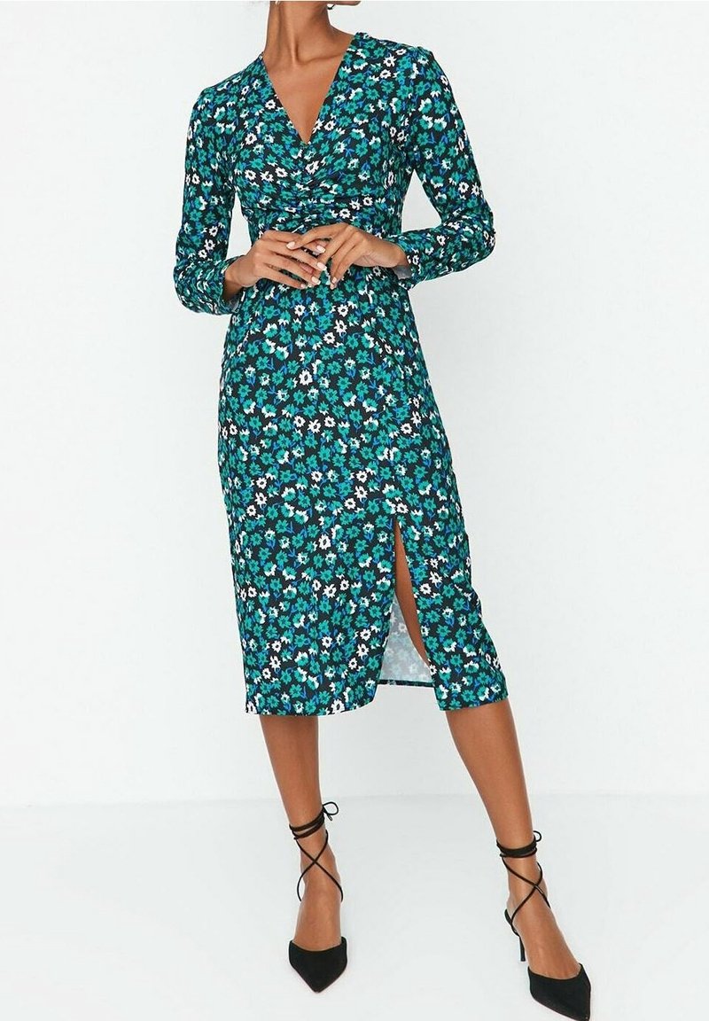 Femme portant une robe portefeuille florale teal à manches longues, avec une fente à l'avant, et des escarpins à bout pointu noirs avec des brides autour de la cheville.