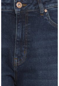 Nærbillede af mørkeblå denimjeans med kobberknap og syningsdetaljer på linningen og gylpen.