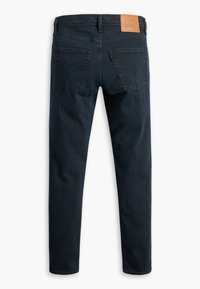 Jean en denim bleu foncé coupe slim, deux poches arrière, une plaque de marque en cuir marron sur la ceinture, et une petite étiquette rouge sur la poche droite.