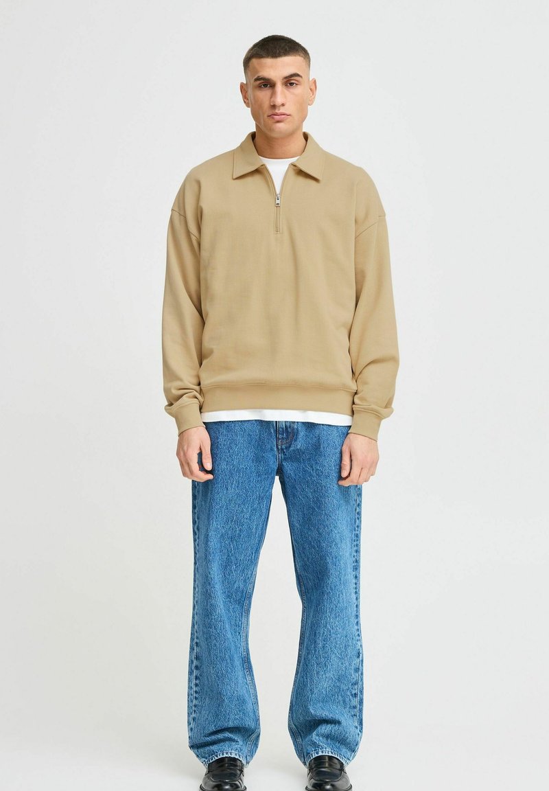 Beige kwart-rits sweatshirt met een relaxte pasvorm, lange mouwen en geribbeld zoom, gecombineerd met lichtblauwe baggy jeans en zwarte schoenen.