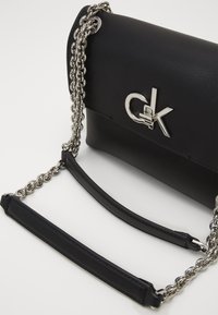 Sort faux læder taske med en sølv kædelsesnor. Har et fremtrædende sølvlogo fortil og en glat, struktureret overflade.