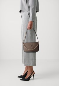 Brun lærveske med kjedestropp, rektangulær form, med merkevaren sin logo. Modellen har på seg en svart og hvit rutete dress og høye hæler.