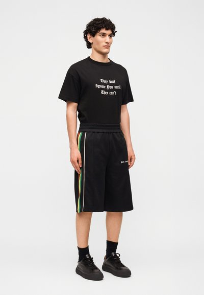 Άνδρας στέκεται φορώντας μαύρο T-shirt με λευκό κείμενο "They will ignore you until they can't", μαύρο σορτς με πράσινες, κίτρινες και κόκκινες ρίγες και μαύρα αθλητικά παπούτσια.