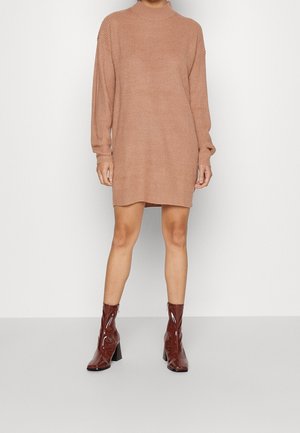 Kobieta ubrana w długą, camelową dzianinową sukienkę z długim rękawem, w połączeniu z błyszczącymi ciemnoczerwonymi botkami, stojąca na tle jednolitego, jasnego tła.