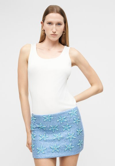 Γυναίκα στέκεται με το ένα χέρι στη μέση, φορώντας άσπρο αμάνικο τοπ και μπλε μίνι φούστα διακοσμημένη με σχέδια αστερία σε ανοιχτό μπλε χρώμα.