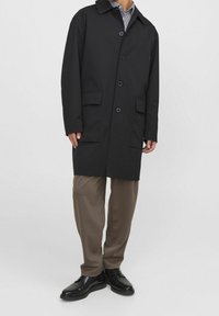 Black coat made of a smooth fabric, featuring a collar, front buttons, and two patch pockets. Worn over brown pants and black shoes.