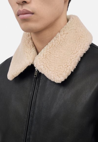 Homme portant une veste en cuir noire avec un grand col en shearling beige et duveteux ainsi qu’une fermeture éclair frontale, visible du menton à la poitrine.