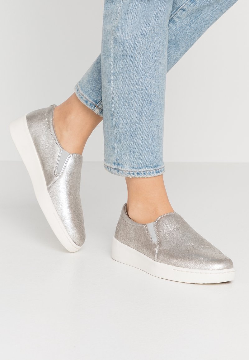 Chaussures en argent métallique à enfiler avec une finition texturée, dotées de panneaux latéraux élastiques et d'une semelle en caoutchouc blanche. Portées avec un jean bleu clair.