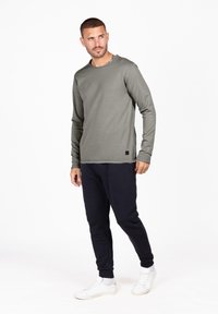 Olivengrünes Langarmshirt mit Rundhalsausschnitt, feinen Säumen und einem schwarzen Logopatch, kombiniert mit marineblauen Hosen und weißen Sneakers.