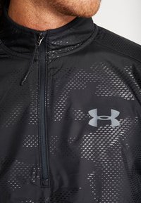 Chaqueta deportiva negra con cremallera media, que presenta un patrón texturizado, detalles reflectantes y un logo gris en el pecho. Acabado de tela suave.