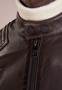 Brun läderjacka med hög krage och en krämfärgad stickad kant, tryckknappar och dragkedja fram. Texturerad yta synlig.