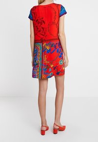 Czerwona sukienka z krótkimi rękawami, ozdobiona mieszanką wzorów kwiatowych i geometrycznych. Materiał wydaje się gładki, dopełniony czerwonymi szpilkami.