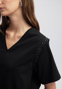 Maglietta nera a collo a V con dettagli texturizzati sulle spalle, piccoli rivetti argentati, maniche corte e in morbido cotone.