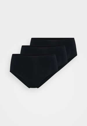 Kvinne iført svarte høywaist truser og en matchende svart crop top med blonder på skulderstroppene, står mot en hvit bakgrunn.