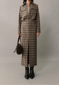 Chemise à carreaux marron superposée sur une jupe ajustée assortie, avec une fente à l'avant. Portée avec des bottines noires à bout pointu et un sac à main marron.