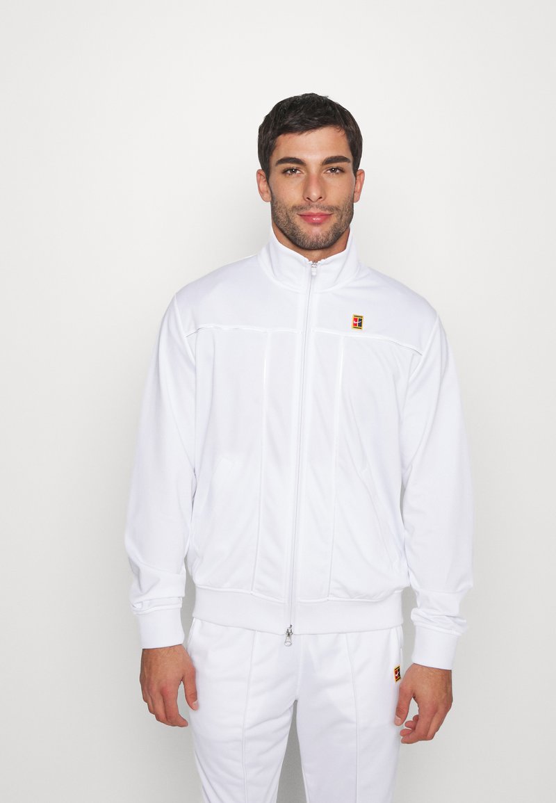 Chaqueta atlética blanca con un alto cuello, cremallera completa y puños acanalados. Presenta costuras sutiles y un parche con el logo en el lado izquierdo del pecho.