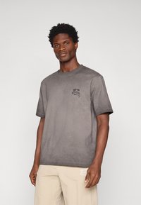 Szary bawełnowy t-shirt z krótkimi r�ękawami, okrągłym dekoltem i minimalistyczną czarną grafiką na lewej stronie klatki piersiowej.