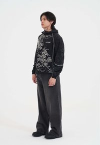 Giacca nera con cappuccio decorata con motivi di drago bianco e nuvole, maniche elastiche, abbinata a pantaloni grigi larghi e scarpe nere con dettagli fiammeggianti.