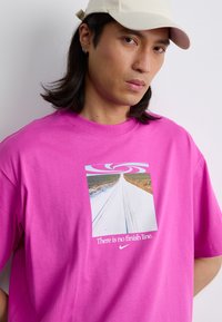 Lyserød bomulds T-shirt med et kvadratisk grafisk design, der viser en hvid vej og et virvlende mønster, med teksten "Der er ingen mållinje" og Nike-logoet.