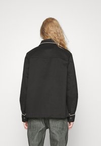 Černá bunda s klasickým límcem, bílým lemováním a zapínacími manžetami. Tkanina vypadá hladce; design je jednoduchý a přiléhavý.