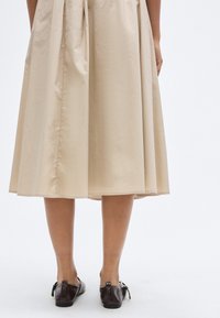 Μπεζ, φούστα μήκους ως το γόνατο με πιέτες από λείο ύφασμα. Συνδυάζεται με σκούρα καφέ, επίπεδα παπούτσια με φιόγκους.