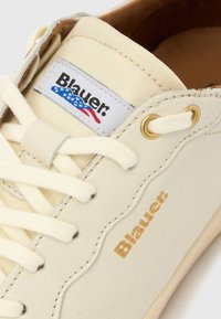 Ténis de cor creme com acabamento em pele texturada, apresentando uma língua em lona branca, ilhós dourados e a marca "Blauer" em dourado.