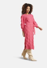 Donna in un vestito midi maglione rosa testurizzato con cintura e stivali beige slouchy con tacco alto, che cammina con una posa sicura su uno sfondo bianco.