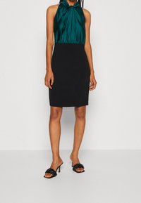 Černé šaty do délky kolen s tyrkysovým saténovým zavazováním za krkem, mají zavinovací korzet. Černé sandály na otevřenou špičku s pletenými popruhy dokončují outfit.