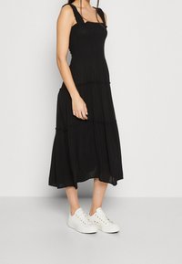 Femme portant une robe midi noire sans manches avec des bretelles froncées et des baskets blanches basses, se tenant devant un fond clair.