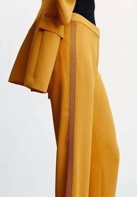 Pantalon jaune moutarde avec bordure décorative sur le côté, porté avec une veste assortie et un haut noir, présenté de la mi-torse à la mi-cuisse sur fond blanc.