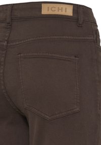 Pantalon marron avec une poche arrière unique et une étiquette beige cousue au-dessus de la taille affichant le nom de la marque ICHI.