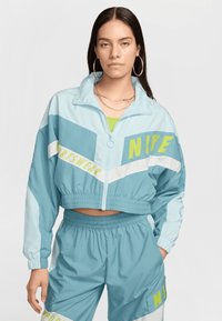 Jachetă scurtă de vânt de culoare albastră deschisă cu accente verzi și albe, având brandingul "Nike", asociată cu pantaloni scurți asortați, ambele dintr-un material neted.