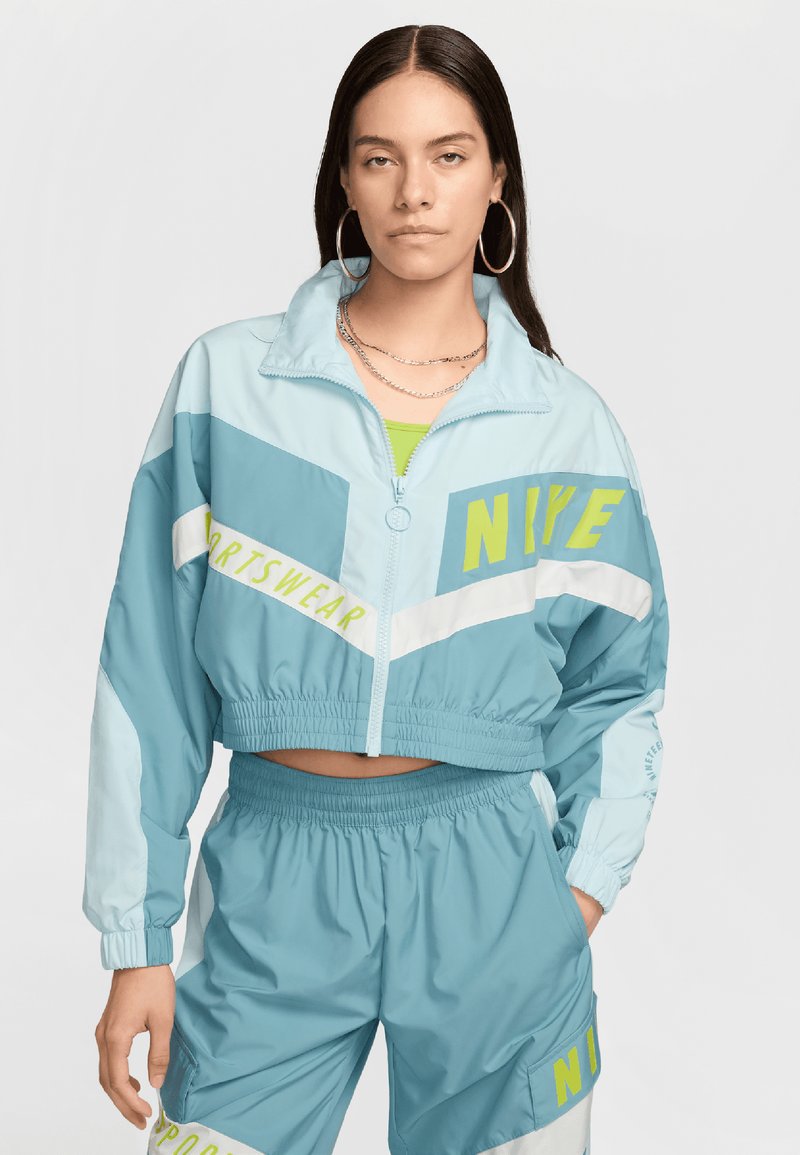 Jachetă scurtă de vânt de culoare albastră deschisă cu accente verzi și albe, având brandingul "Nike", asociată cu pantaloni scurți asortați, ambele dintr-un material neted.