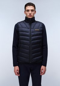 Chaleco de pluma negro con cuello alto, patrón de acolchado vertical, cremallera frontal y un pequeño parche de logo en el pecho izquierdo. Lleva puesto sobre ropa oscura.