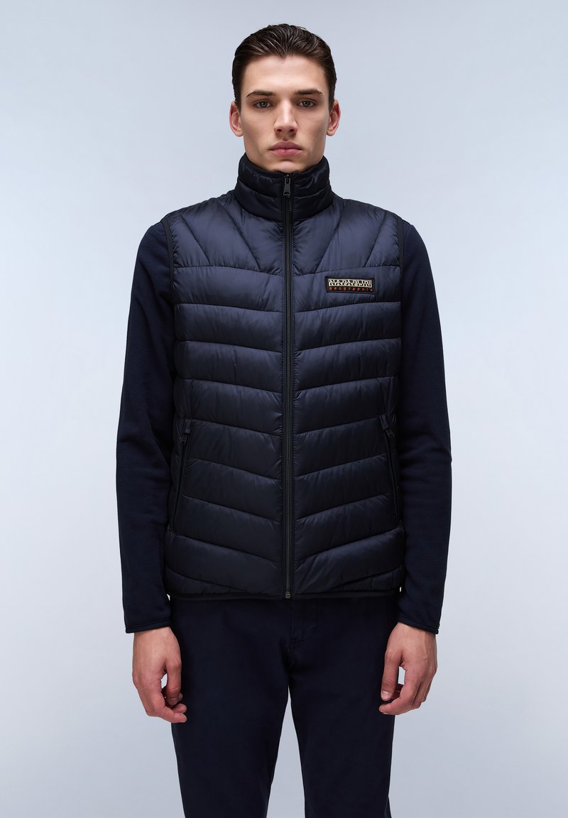 Chaleco de pluma negro con cuello alto, patrón de acolchado vertical, cremallera frontal y un pequeño parche de logo en el pecho izquierdo. Lleva puesto sobre ropa oscura.