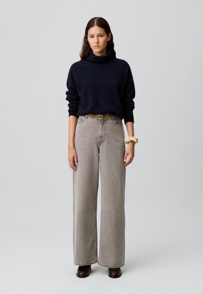 Mujer con cabello largo y oscuro que lleva un suéter negro de cuello alto, jeans grises de pierna ancha con cinturón y zapatos marrones, de pie contra un fondo sencillo.