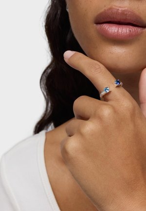 Anillos de plata esterlina con una piedra preciosa azul claro y otra azul oscuro. La mano está colocada de manera reflexiva cerca de los labios.